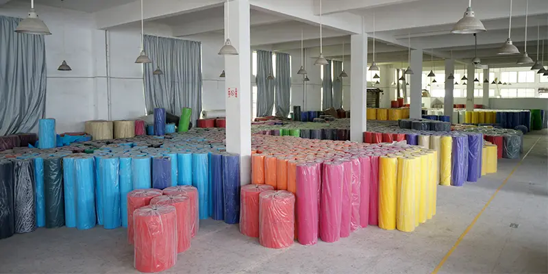 Rows of colorful non-woven fabric rolls neatly stacked inside a factory warehouse, showing different thicknesses and material quantities.