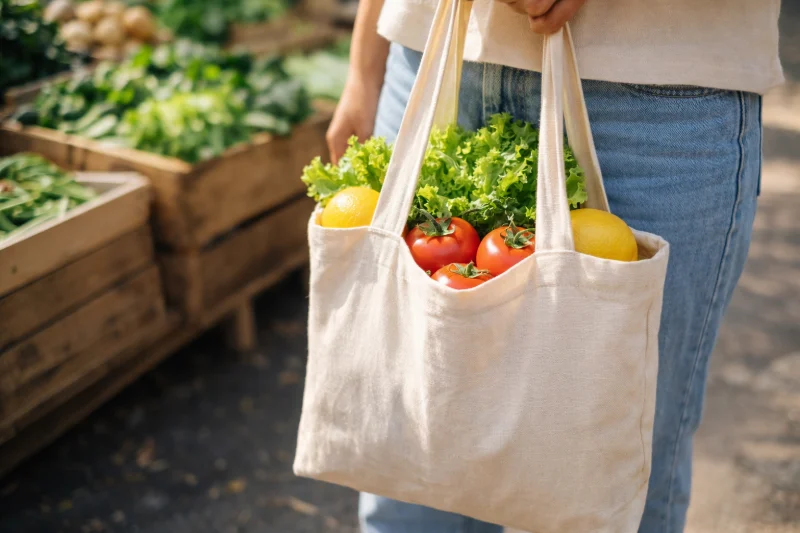 A cotton fabric eco bag filled with fresh vegetables and fruits being carried at an outdoor market, showing what is cotton fabric explained for eco bag buyers.