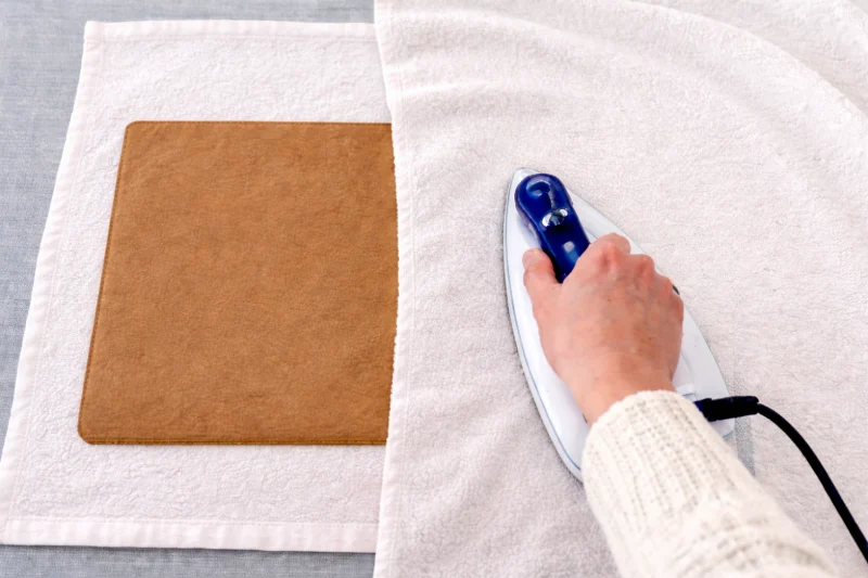 A washable kraft paper bag lies flat on a towel while a hand uses an iron nearby to help smooth and dry it.
