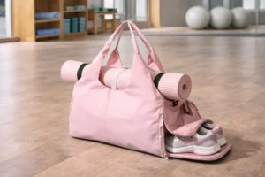A pink gym bag style sits on a wooden studio floor, holding a rolled yoga mat on top and athletic shoes stored in a side compartment.