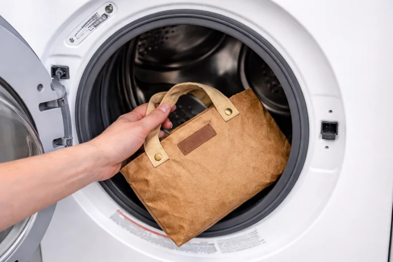 A hand placing a washable kraft paper bag inside a washing machine drum for cleaning.