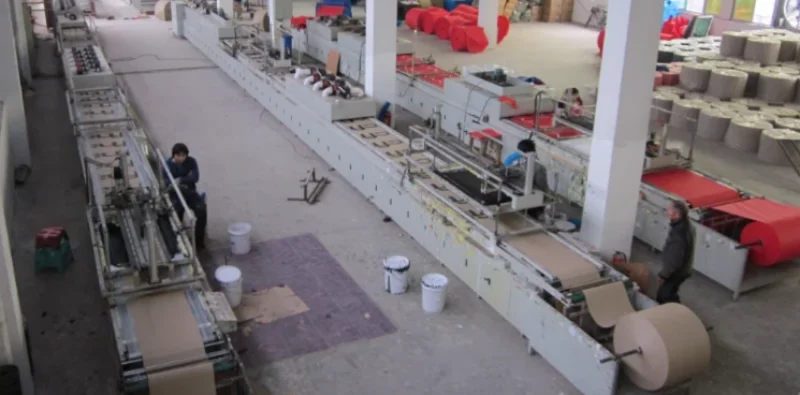 An automated material processing line runs inside a large factory hall with machinery and organized workstations.