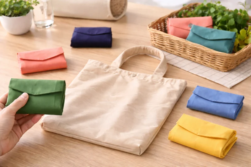 A beige foldable bag laid flat on a wooden table surrounded by neatly folded compact pouches in various colors, illustrating Your Complete Guide to Foldable Bags.