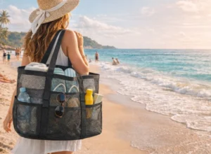 A person wearing a sun hat walks along the shoreline carrying a large mesh beach tote filled with beach items, illustrating Best Materials for Beach Bags.