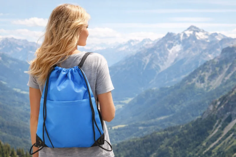 A person wearing a blue drawstring backpack outdoors in a mountain setting, demonstrating what is polyester fabric and why is it used for reusable shopping bags.