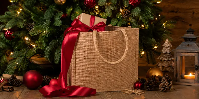 A jute gift bag with a wrapped present is placed in a Christmas decorated setting for holiday packaging.